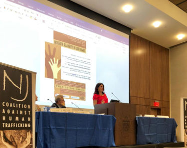 A woman speaking at a podium with a slideshow behind her at the Modern Slavery Symposium.
